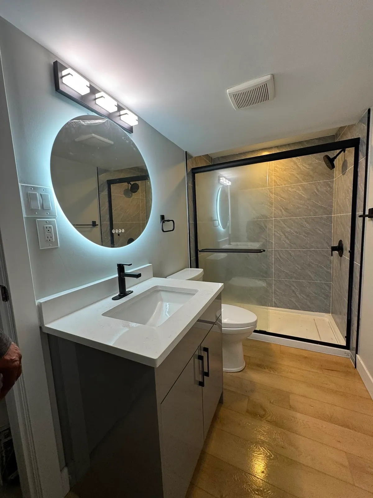 Same bathroom after renovation with backlit round mirror, black fixtures, and glass shower